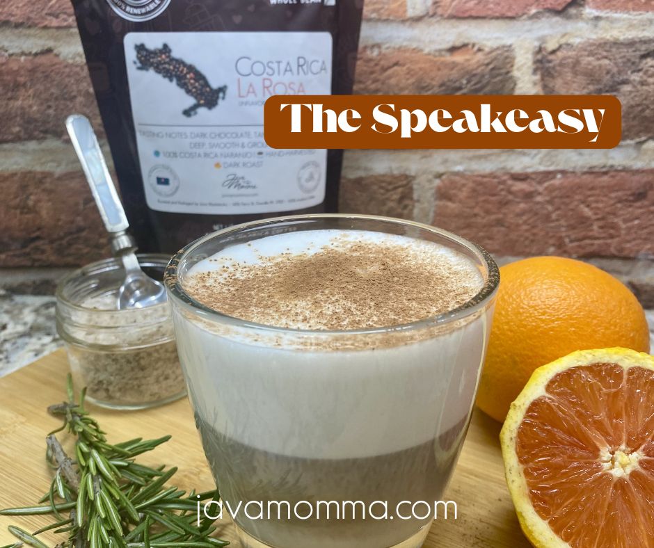 A layered latte called The Speakeasy, topped with cocoa powder and frothed milk, surrounded by rosemary sprigs, a halved orange, smoked salt, and a bag of Costa Rica La Rosa coffee beans against a rustic brick background.