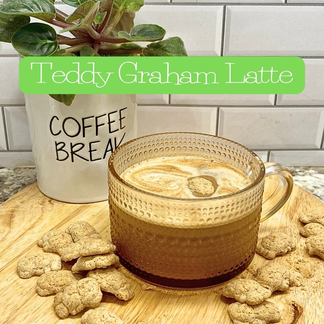 A glass mug of Teddy Graham Latte topped with frothy milk and a small Teddy Graham cookie floating on top. The mug sits on a wooden board surrounded by Teddy Grahams, with a white “Coffee Break” mug and a green plant in the background.