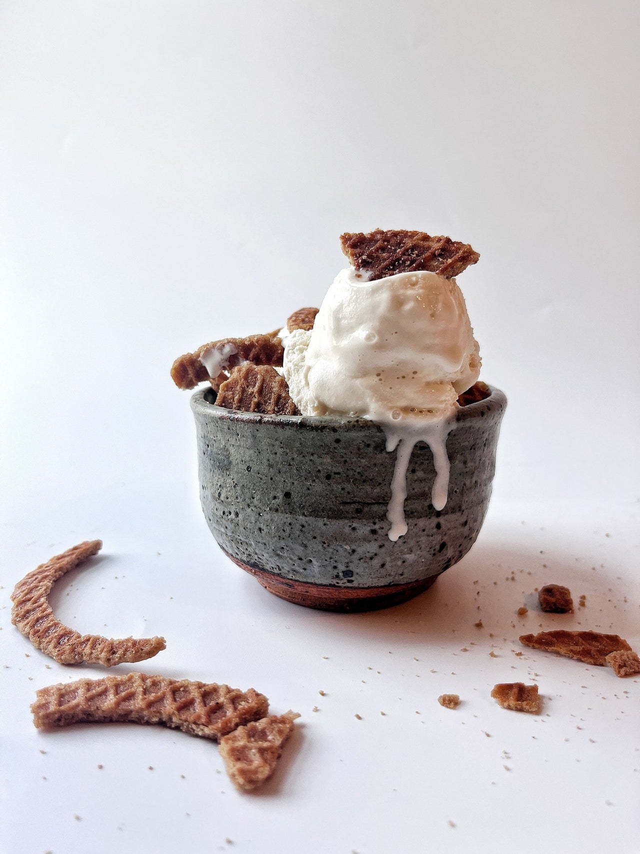 A scoop of vanilla ice cream sits in a gray bowl, topped and surrounded by pieces of Original Plain Stroopie Singles – Classic Caramel Comfort, with extra cookie crumbs scattered on a white surface.