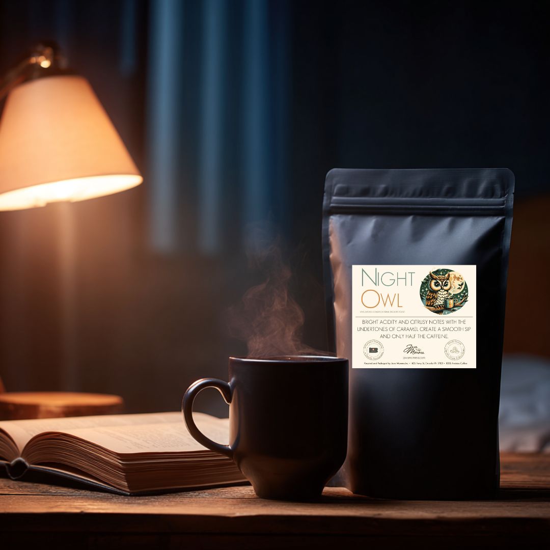 Java Momma Night Owl Half Caff coffee bag on a wooden table beside a steaming mug of coffee and an open book, warm evening lamp light creating a cozy nighttime scene.