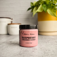 Thumbnail for A jar of Nate’s Nectar Raspberry Creamed Honey – Tangy, Sweet & Spreadably Smooth sits on a gray countertop with kitchen canisters and a green potted plant in the background, its label clearly visible.