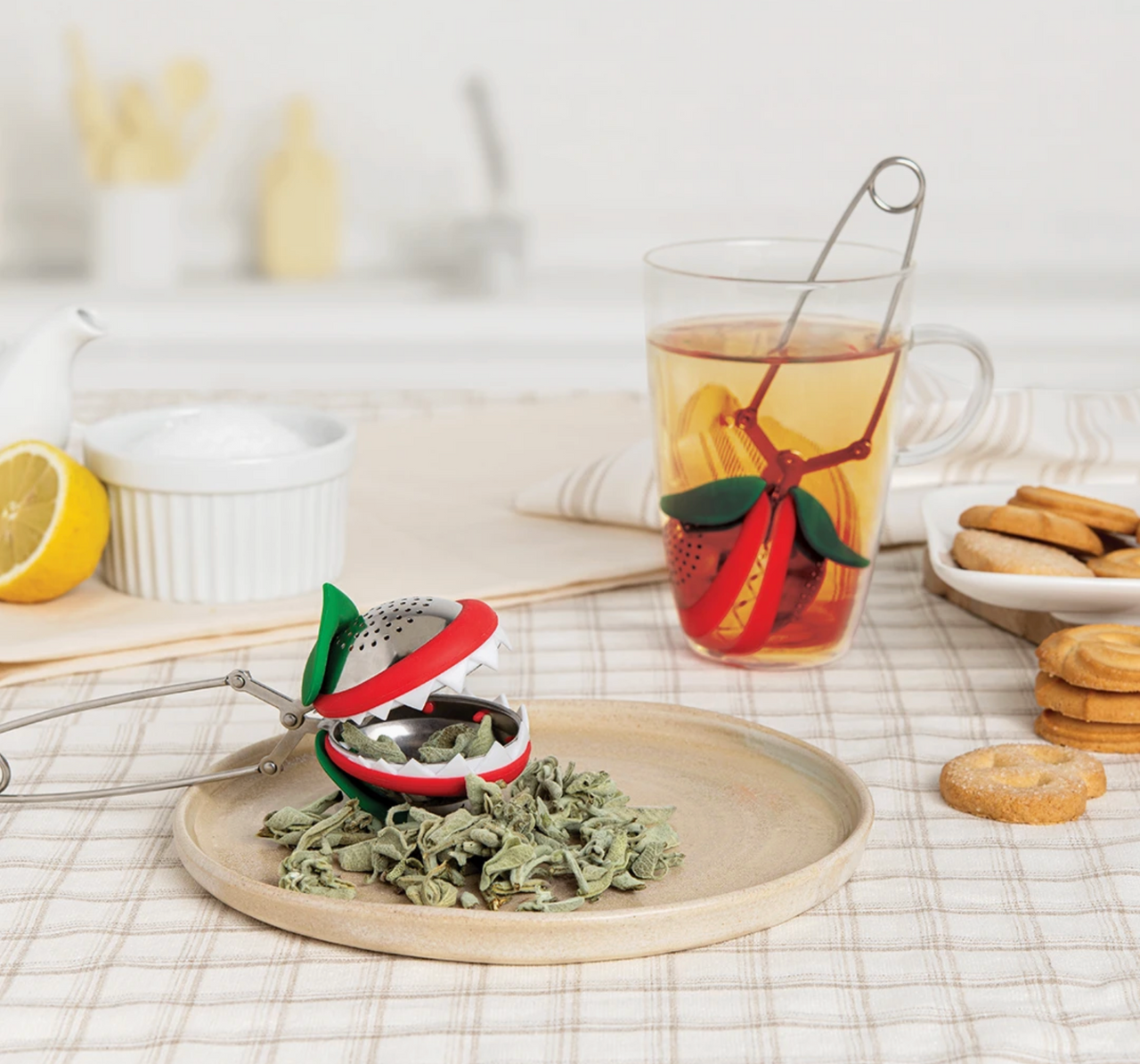 The 🪴 Steep Fiend – The Loose Leaf Monster That Lives to Brew by OTOTO Studio, shaped like a plant, sits with loose leaf tea on a plate as another infuser steeps in a glass; cookies, lemon, sugar, and milk are nearby.