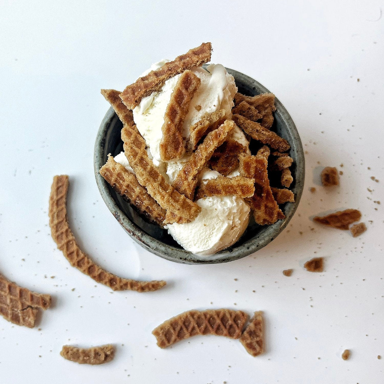 A bowl of vanilla ice cream topped with Stroopwafel Just the Cookies 🧇 – 100g Bag of Sweet Snacking Bliss, with extra waffle cookie pieces scattered around the bowl on a white surface.