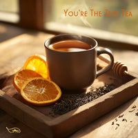 Thumbnail for A sleek ceramic mug filled with rich amber-colored You're the Zest black tea, placed on a rustic wooden tray. Fresh orange slices, dried black tea leaves, and a honey dipper surround the cup, creating a warm and invigorating scene. Soft golden sunlight filters through a nearby window, enhancing the cozy yet refreshing citrus-inspired atmosphere.