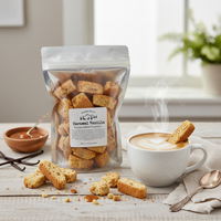 Thumbnail for A bag of Caramel Vanilla Biscotti Bites sits on a wooden table beside a steaming cappuccino with latte art. Several dunkable bites are scattered around, and one rests on the rim of the cup.