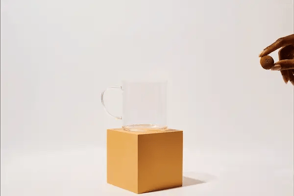 A hand drops a Pumpkin Spice Tea Drop – Cozy Up to Fall in a Cup 🎃☕🍂 into a clear glass mug on a yellow cube, creating an orange crown-shaped splash. The background is simple and light-colored.