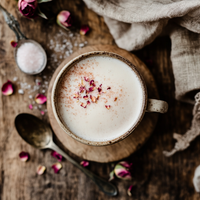 Thumbnail for A ceramic mug of frothy milk topped with pink rose petals and a sprinkle of 🌹 Rose Petal Sugar Sprinkle sits on a wooden surface, surrounded by a spoon, linen napkin, dried roses, and coarse salt.