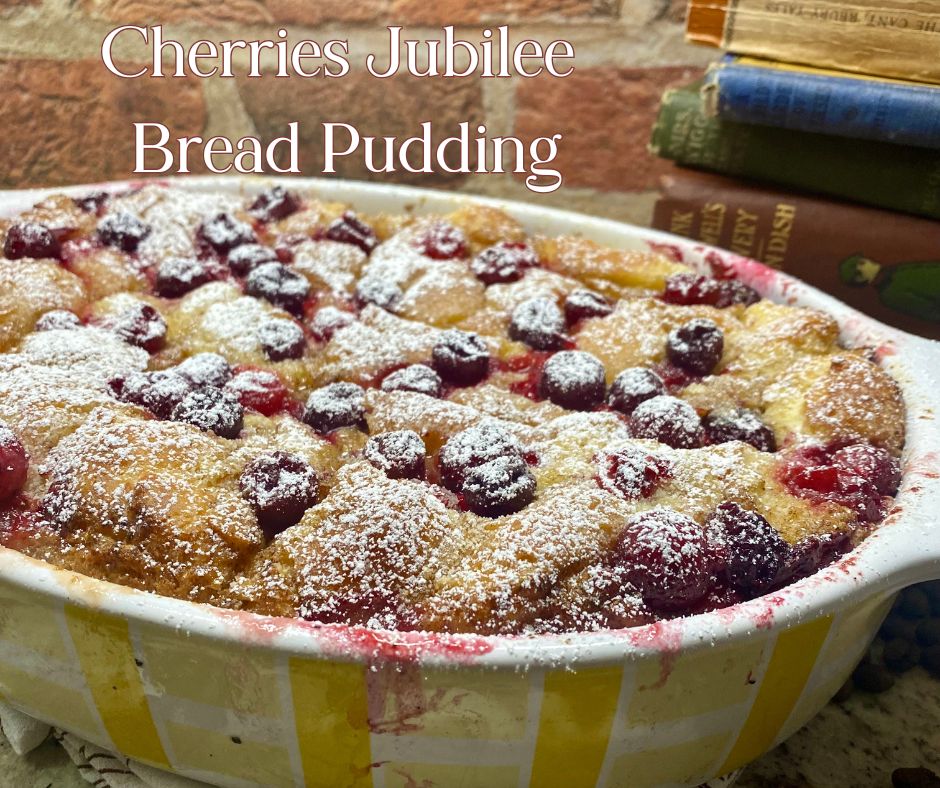 A golden-brown Cherries Jubilee Bread Pudding in a yellow checkered baking dish, topped with plump cherries and dusted with powdered sugar. The dish is set against a rustic brick background with stacked vintage books beside it.