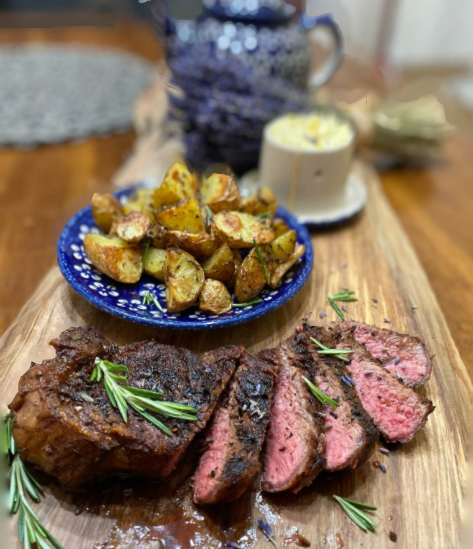 Sliced Lavender-Rosemary Steak served on a wooden board with sprigs of fresh rosemary, alongside a blue patterned plate of golden roasted potatoes and a ramekin of whipped lavender honey butter. A teapot and herbs are softly blurred in the background.