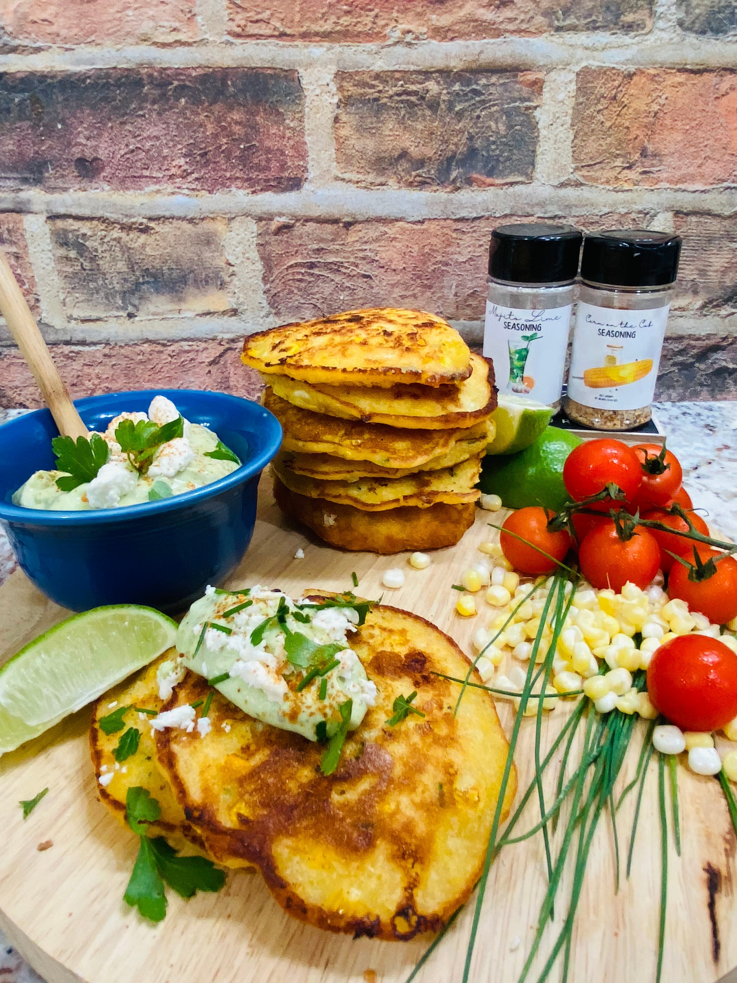 Golden sweet corn fritters stacked on a plate with a dollop of green avocado crema, garnished with fresh chives and surrounded by lime wedges and corn kernels.