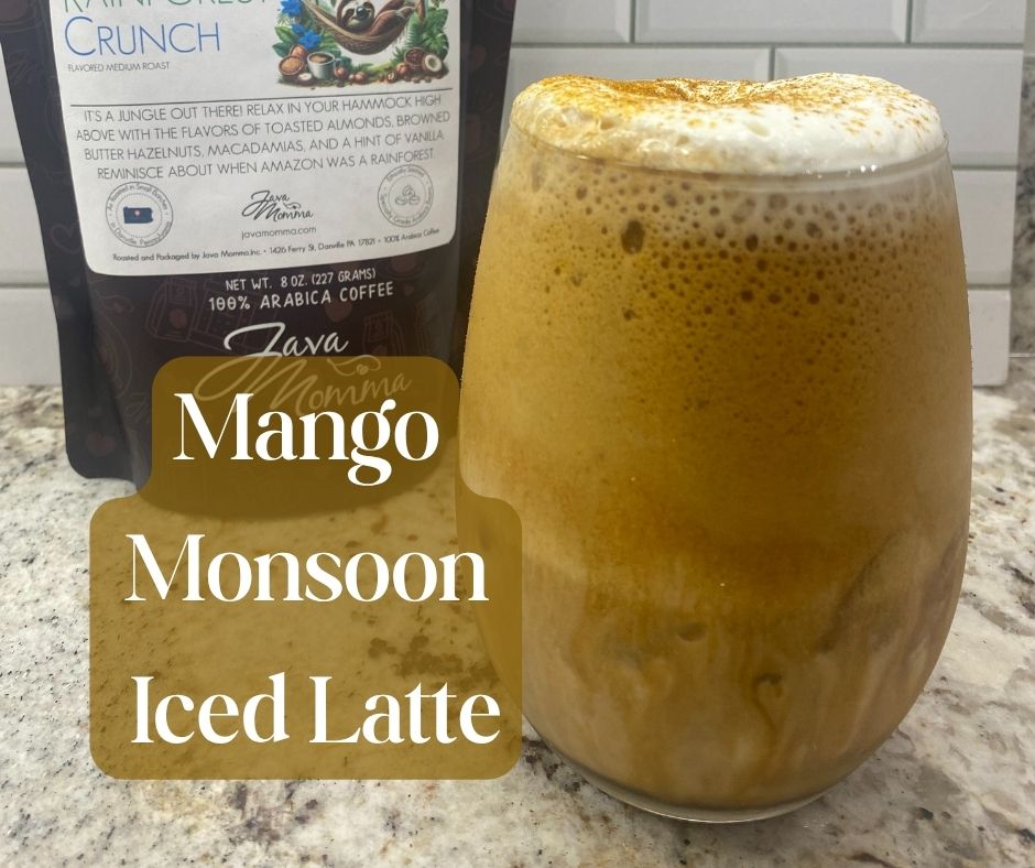 A glass of iced Mango Monsoon espresso drink topped with frothy foam and cinnamon, placed in front of a bag of Java Momma Rainforest Crunch whole bean coffee on a granite countertop with a white subway tile backsplash.