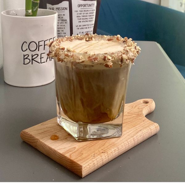 A Frosted Sticky Bun Latte served in a square glass, topped with frothy milk and drizzled caramel, with the rim coated in crushed pecans. The drink sits on a small wooden board, styled beside a white coffee mug labeled “Coffee Break.