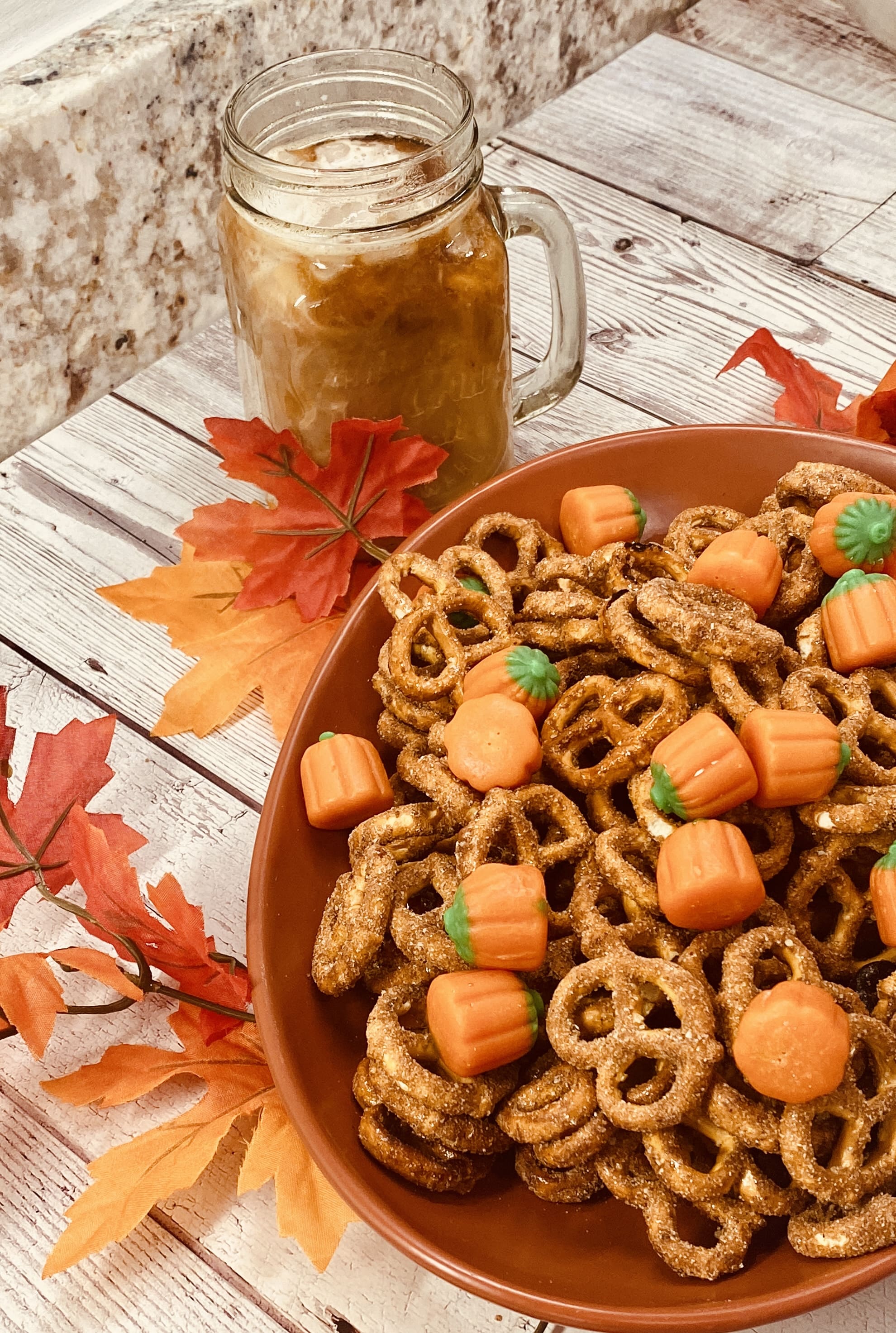 Bowl of sweet and salty pretzel twists coated in pumpkin spice sugar, mixed with soft pumpkin mallows, surrounded by fall leaves and a mason jar drink.