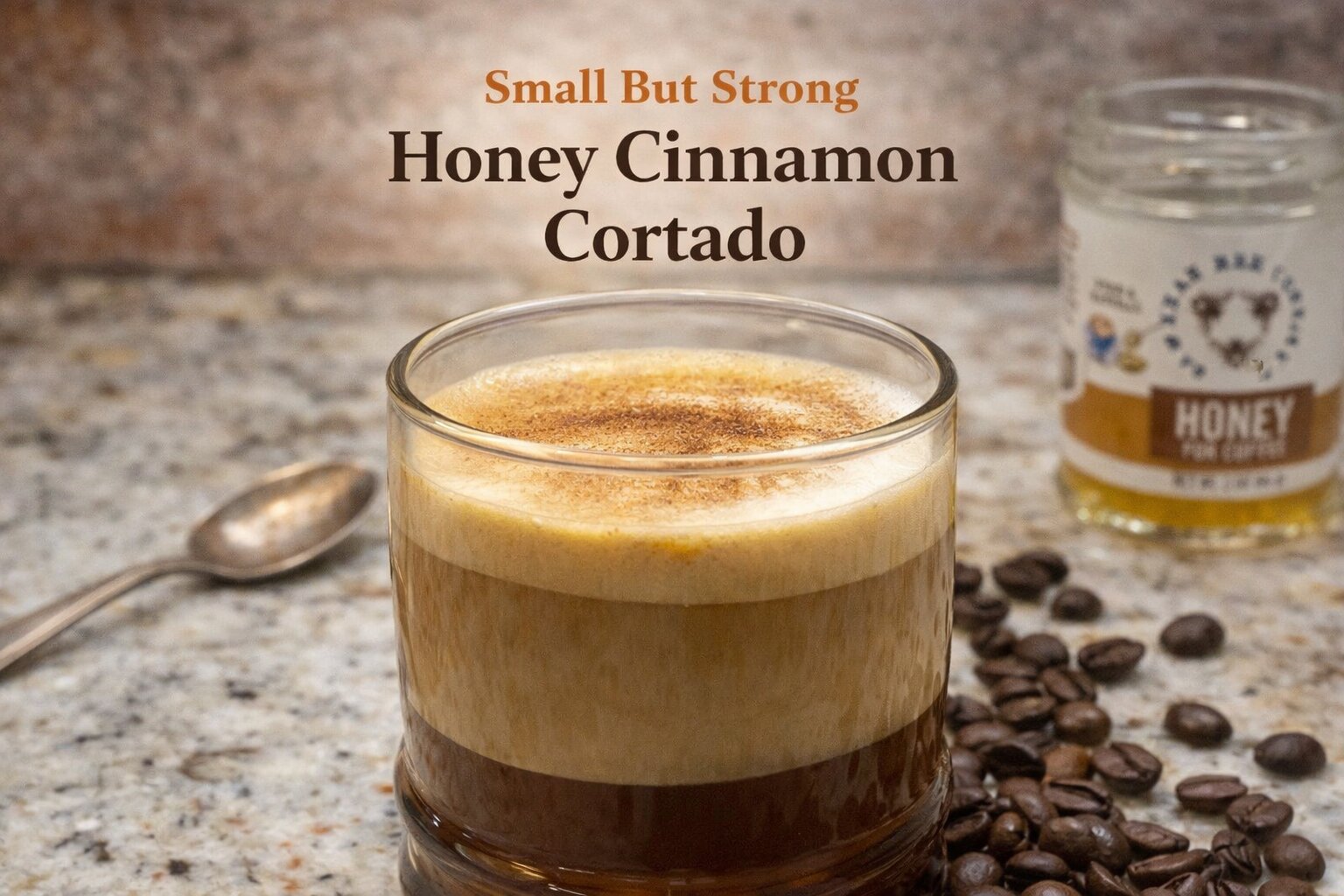 Glass of honey cinnamon cortado with layered espresso and steamed milk on a granite countertop, coffee beans scattered nearby and honey jar in the background.