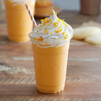 Thumbnail for A clear cup of 🍊 Orange Creamsicle Frappe Mix – Creamy, Dreamy, and Totally Nostalgic sits on a wooden table, topped with swirled whipped cream and orange zest, striped straw inserted, with blurred baked treats in the background.