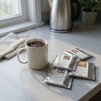 Thumbnail for A ceramic mug of hot mocha sits on a clean kitchen counter beside four loosely grouped Java Momma drink mix packets, with a kettle and plant visible in the background.