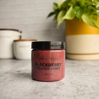 Thumbnail for A jar of Nate’s Nectar Blackberry Creamed Honey – Berry Sweet & Spreadably Smooth rests on a gray countertop, with a plant in a yellow pot and two white canisters softly blurred in the background.