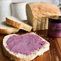 Thumbnail for A slice of white bread spread with Nate’s Nectar Blueberry Creamed Honey 💙 rests on a wooden board beside a loaf, two slices, and the jar. White canisters blur in the background.