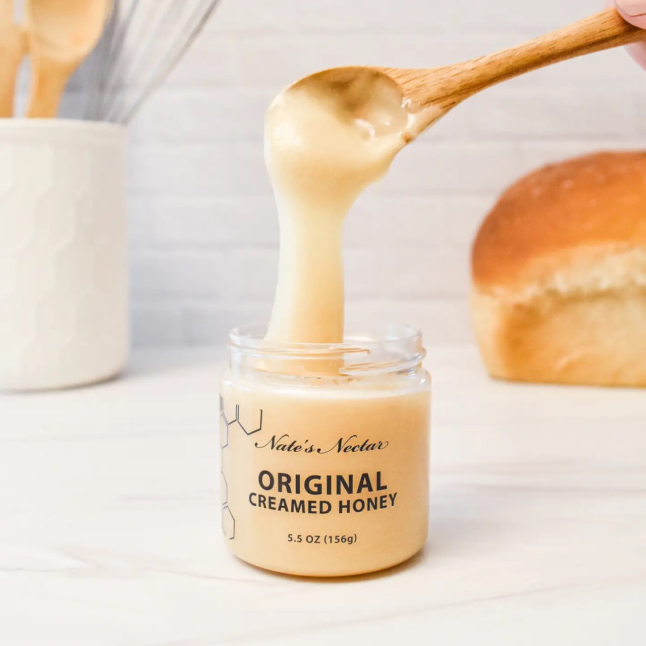 A wooden spoon dips into a jar of Nate’s Nectar Original Creamed Honey 🐝 – Pure, Spreadable Bliss in a Jar, lifting out smooth honey; a loaf of bread and kitchen utensils are softly blurred in the background.