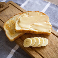 Thumbnail for Two slices of bread, one plain and one topped with Nate’s Nectar Original Creamed Honey 🐝, rest on a wooden board with five banana slices in front. The board is placed on a striped cloth.