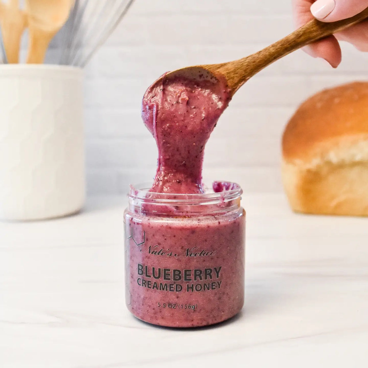 A wooden spoon lifts thick, purple Nate’s Nectar Blueberry Creamed Honey 💙 – A Berry-licious Spread You’ll Crave from a glass jar. A loaf of bread and kitchen utensils in the background highlight this rich, raw honey treat.