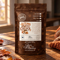 Thumbnail for A stand-up pouch of Java Momma 'Autumn Toffee Crunch' coffee stands on a rustic wooden table with scattered autumn leaves and a knitted orange-brown scarf in the background. The package is dark brown with coffee-themed illustrations and a large white front label.