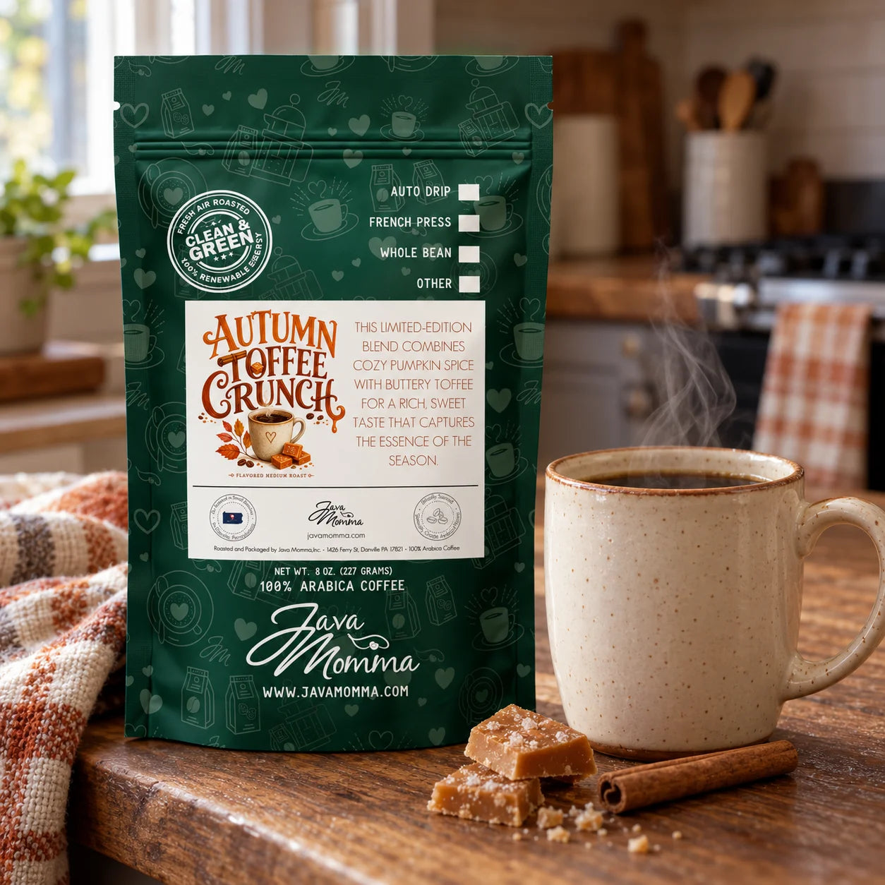 A bag of Java Momma 'Autumn Coffee Crunch' coffee stands on a wooden table next to a steaming ceramic mug, with cinnamon and sugar nearby and a softly blurred kitchen background.