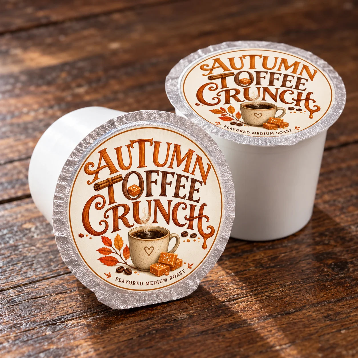 Two single-serve coffee pods labeled 'Autumn Toffee Crunch' on a rustic wooden surface, with one pod shown open to reveal the white coffee cup inside and the other closed, displaying the branded foil lid.