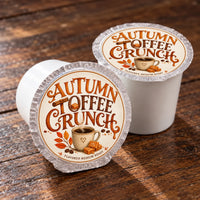 Thumbnail for Two single-serve coffee pods labeled 'Autumn Toffee Crunch' on a rustic wooden surface, with one pod shown open to reveal the white coffee cup inside and the other closed, displaying the branded foil lid.