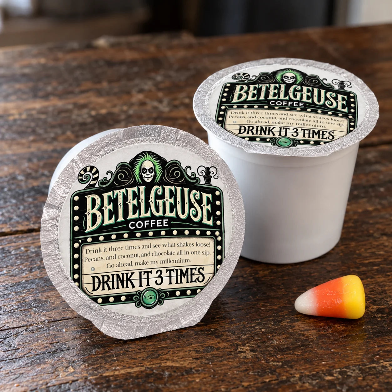 Two Betelgeuse Coffee K-cups on a rustic wooden table, with one cup open and one sealed, alongside a candy corn piece. The labels feature a gothic green and black design with the text 'BETELGEUSE COFFEE' and 'DRINK IT 3 TIMES'.