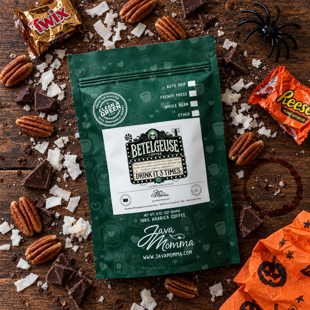 A green stand-up coffee pouch labeled 'Bettelgeuse' and 'Java Momma' is arranged on a rustic wooden surface with Halloween-themed candy, pecans, chocolate pieces, and orange paper. The product is the coffee pouch, photographed as a styled flat-lay.