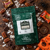 Thumbnail for A green stand-up coffee pouch labeled 'Bettelgeuse' and 'Java Momma' is arranged on a rustic wooden surface with Halloween-themed candy, pecans, chocolate pieces, and orange paper. The product is the coffee pouch, photographed as a styled flat-lay.
