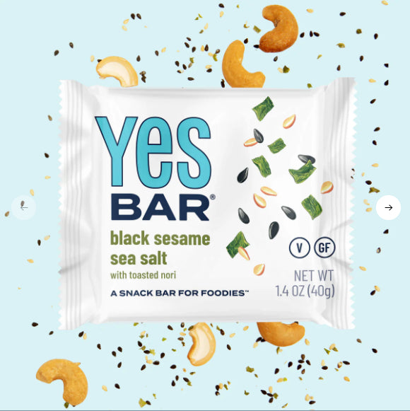 A package of Black Sesame Sea Salt YES Bar® – Sweet, Savory, and Totally Addictive sits on a light blue background with cashews, sesame seeds, and seaweed; this 1.4 oz plant-based snack features toasted nori for a savory twist.