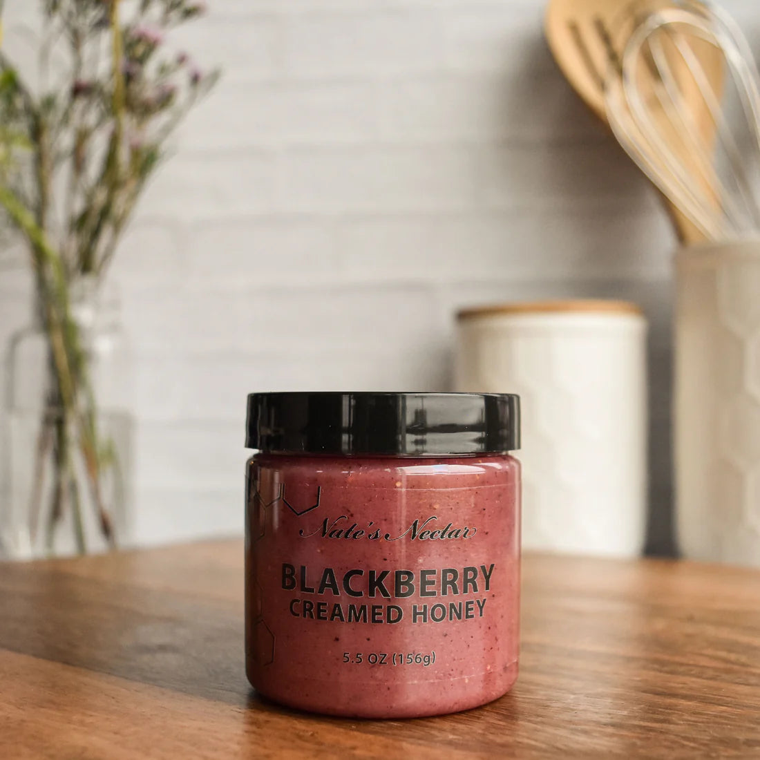 A jar of Nate’s Nectar Blackberry Creamed Honey – Berry Sweet & Spreadably Smooth sits on a wooden countertop, with kitchen utensils in a white holder and a blurred flower bouquet creating a cozy background.