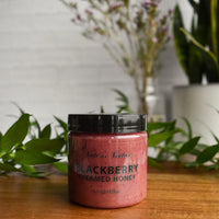 Thumbnail for A jar of Nate’s Nectar Blackberry Creamed Honey – Berry Sweet & Spreadably Smooth rests on a wooden surface with green leaves and blurred floral vases in the background, showcasing its rich berry flavor and smooth texture.