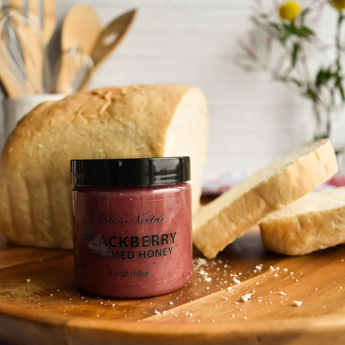 A jar of Nate’s Nectar Blackberry Creamed Honey – Berry Sweet & Spreadably Smooth sits before sliced bread on a wooden board, with blurred kitchen utensils and flowers in the background, emphasizing its rich, spreadable flavor.