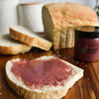 Thumbnail for A slice of white bread topped with Nate’s Nectar Blackberry Creamed Honey sits on a wooden surface, with a loaf, extra slices, and the creamed honey jar in the background.