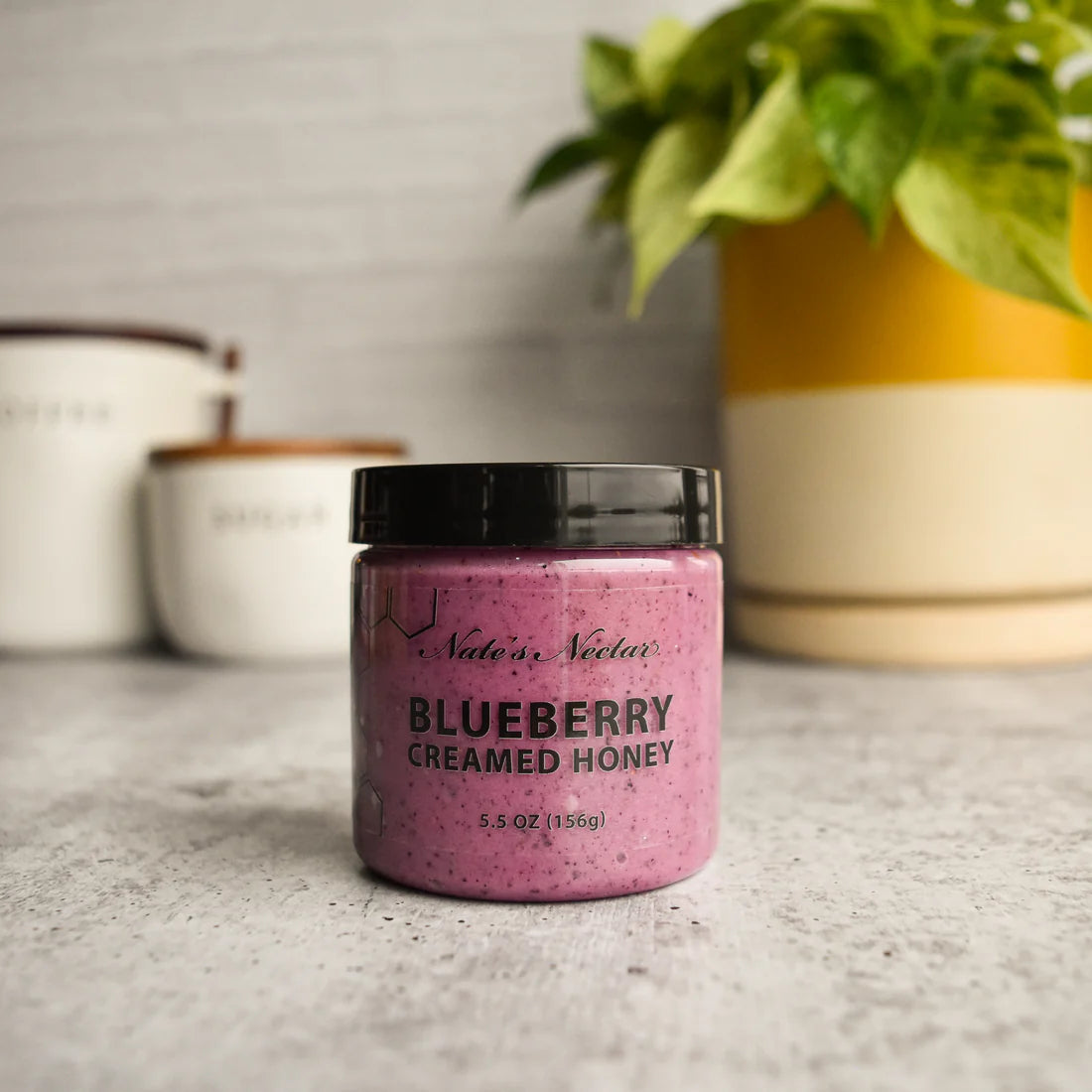 A jar of Nate’s Nectar Blueberry Creamed Honey 💙 – A Berry-licious Spread You’ll Crave sits on a gray countertop, with blurred white canisters and a green potted plant in the background.