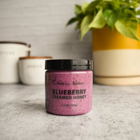 Thumbnail for A jar of Nate’s Nectar Blueberry Creamed Honey 💙 – A Berry-licious Spread You’ll Crave sits on a gray countertop, with blurred white canisters and a green potted plant in the background.