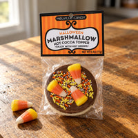 Thumbnail for A packaged round marshmallow topped with chocolate, candy corn, and sprinkles, hanging from an orange Melville Candy Halloween Marshmallow Hot Cocoa Topper card, on a wooden table with two candy corn pieces beside it.