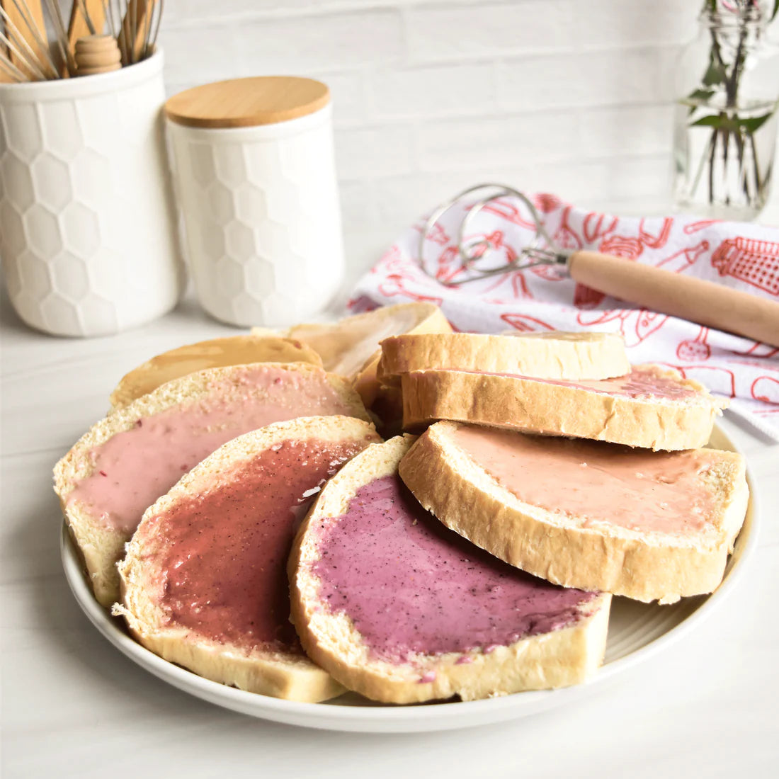 Slices of white bread spread with Nate’s Nectar Peach 🍑 Creamed Honey sit on a plate. Kitchen canisters, utensils, and a floral cloth are seen in the background on a white countertop.
