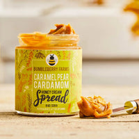 Thumbnail for A jar of Bumbleberry Farms Caramel Pear Cardamom Honey Cream Spread sits on a wooden surface with a dollop of the spread and a small spoon visible. The jar has a yellow-green label with botanical illustrations and the product name in large text.