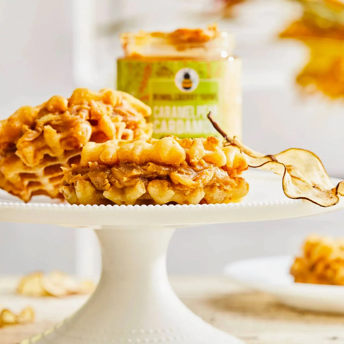 Caramelized cardamom cookies on a white pedestal plate with a jar of peanut butter and caramel sauce in the background.