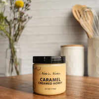 Thumbnail for A jar of Nate’s Nectar Caramel Creamed Honey 🍯 – Sweet, Buttery Bliss in a Jar sits on a wooden countertop among kitchen utensils, a honeycomb-patterned canister, and flowers—perfect for fans of caramel spread or raw natural honey.