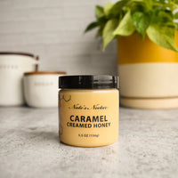Thumbnail for A jar of Nate’s Nectar Caramel Creamed Honey 🍯 – Sweet, Buttery Bliss in a Jar sits on a gray countertop, its rich caramel hue enticing, with a green potted plant and two blurred labeled containers in the background.