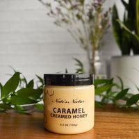 Thumbnail for A 5.5 oz (156g) jar of Nate’s Nectar Caramel Creamed Honey 🍯 – Sweet, Buttery Bliss in a Jar sits on wood, displaying rich caramel creamed honey made from raw natural honey. Green leaves and blurred plants appear in the background.