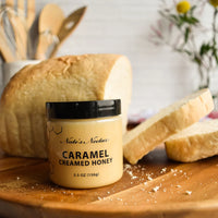 Thumbnail for A jar of Nate’s Nectar Caramel Creamed Honey 🍯 – Sweet, Buttery Bliss in a Jar sits on a wooden surface beside sliced bread, with blurred kitchen utensils and flowers in the background.