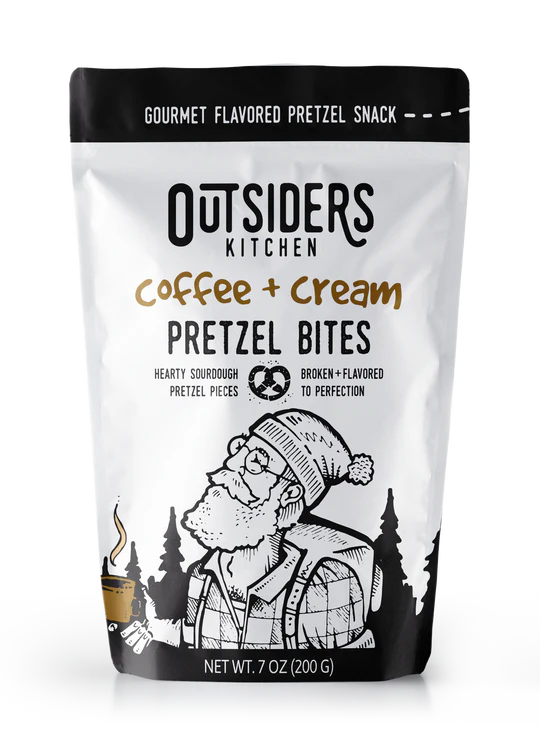 A white bag labeled Coffee + Cream Pretzel Bites – Sweet, Salty, and Seriously Addictive ☕✨, shows a bearded person with a hat and mug in a forest. Enjoy small batch snacks with rich coffee flavor. Net weight: 7 oz (200 g).