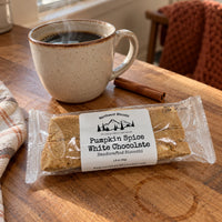 Thumbnail for A packaged bar of Northwest Biscotti Pumpkin Spice White Chocolate Handcrafted Biscotti sits on a wooden table next to a steaming mug of coffee, a cinnamon stick, and a cozy knit blanket.