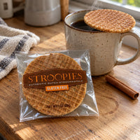 Thumbnail for A packaged round Dutch stroopwafel sits on a wooden table beside a mug of coffee with another waffle resting on the rim, plus a cinnamon stick and cloth in a real indoor setting.